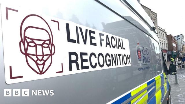 A view taken to show the side of a facial recognition police van. Closest to the camera is a stylised graphic of a face being scanned, next to the text 'Live Facial Recognition'. Further along the van, only partially visible, is the logo for Essex Police
