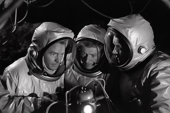Three men in radiation suits huddle in fear around a lamp