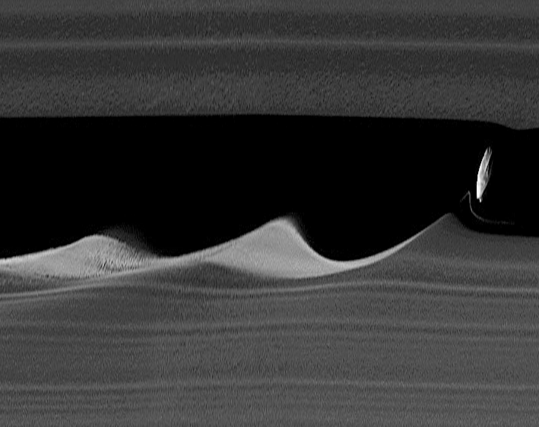A close-up photo of Saturn's ring. The small moon Daphnis appears as an egg-shaped rock inside a cleared band. (The Keeler Gap.) It leaves wave-like patterns in its wake both the inside and outside border of the gap. The photo has been processed (stretched).