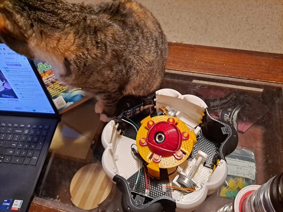 A 3d-printed device that looks like an eyeball with a camera in it surrounded by a casing that contains motors and circuitry, sitting on a coffee table. A tricolor tabby cat, a laptop, and an Asimov's magazine are also visible; the cat is sitting right next to the eyeball like she owns it.