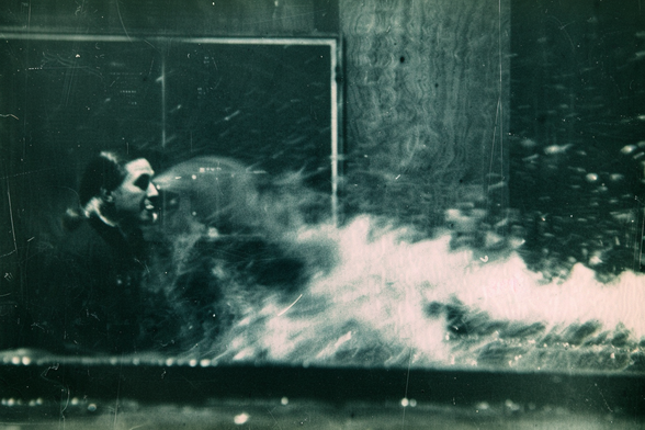 A medium sits at a table in front of a blackboard. Smoke or luminous material covers the table