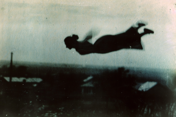 A silhouette of a woman, wearing a dress and her hair in a bun, flying high over a factory
