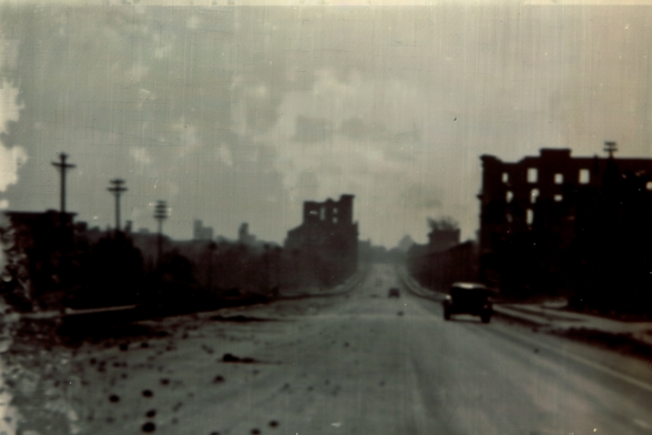 A car drives down a wide but partly rubble-strewn road passing between ruined multi-story buildings