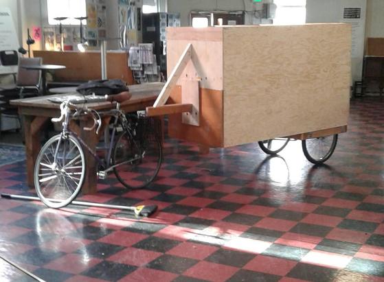 An extremely large wooden box/trailer supported by wheels toward the rear and by a 2x4 tongue hitch resting on the rear rack of a bicycle stands in a large room with black and red tile floor.
