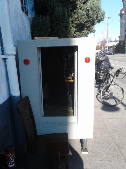 A green-grey box with an open door in the center and red bicycle reflectors on each side.  This is the rear view of a bike camper parked next to a building in Oakland, CA.