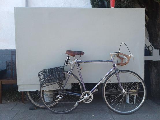 A purple Purple Peugeot road bike with a slanted rear cargo rack is leaned against the side of a pastel green/grey trailer which dwarfs the bicycle.