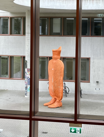 A huge orange hot water bottle standing on its own feet between two buildings at KIT Karlsruhe.