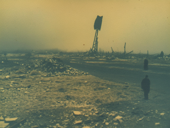 Desolate landscape with debris, a large damaged structure, and two dark figures under a yellowish sky.