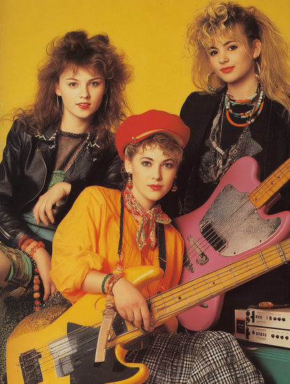 The image features a trio of women posed against a bright yellow background. Each woman holds or is positioned with a musical instrument. The woman on the left wears a black leather jacket with a textured top underneath, accessorized with several colorful bracelets. Her long, light brown hair is styled in a voluminous manner. The central figure sits slightly forward, dressed in a vibrant yellow top with a patterned scarf around her neck and a red beret on her head. She holds a yellow electric bass guitar, her fingers gently resting on the strings. The woman on the right wears a black outfit with layered necklaces and large hoop earrings, her blonde hair is styled with curls and volume. She holds a pink electric bass guitar.