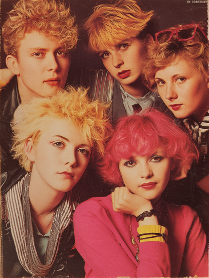 The image is a portrait of five individuals with distinct 1980s fashion and hairstyles. Each person has a unique hairstyle, characterized by bold, voluminous looks and vibrant colors. The person in the center foreground has bright pink hair and is wearing a pink top with layered necklaces and bracelets. To the left, another individual with light blonde hair is dressed in a black and white striped garment. Behind, three other individuals have different shades of blonde hair, styled with volume and texture. They are wearing various jackets and shirts, adding to the eclectic mix of patterns and layers typical of 1980s fashion. The background is a muted brown, highlighting the colorful clothing and hairstyles.

