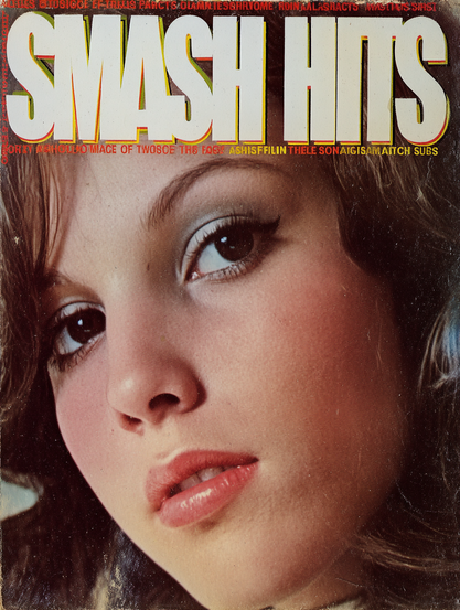 The image is a close-up of a magazine cover titled "Smash Hits." The main focus is a young person's face occupying the lower half of the cover. The face is angled slightly to the left, with full lips and large, expressive eyes accented by makeup. The background is blurred, emphasizing the subject's facial features. The magazine title, "SMASH HITS," is prominently displayed at the top in large, bold white letters with a yellow outline. Below the title, there is a strip of smaller red text.