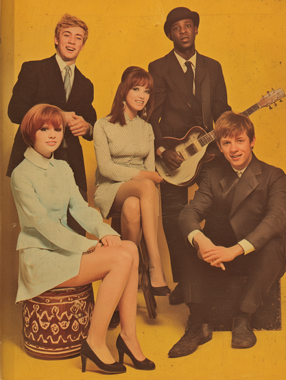 The image features a group of five people, three men, and two women, posing against a bright yellow background. The person on the left stands behind the others, wearing a dark suit with a light shirt and tie, and sporting short blonde hair, smiling pleasantly. In front of him sits a woman with short red hair wearing a light blue outfit and black heels, seated on a decorative cylindrical stool. To her right is another woman with long brown hair, dressed in a light blue patterned dress and black heels, sitting on a chair. Behind her stands a man with a dark suit and tie, wearing a black hat, holding an electric guitar. Finally, to the right is a man seated on a black box, wearing a dark suit with a light shirt, smiling at the camera.