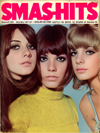 The image is a magazine cover featuring three young women positioned closely together. Each woman gazes directly at the viewer with neutral expressions. The woman on the left has long, straight brown hair and is wearing eyeliner. The middle woman has a blunt, dark brown bob hairstyle and is wearing a bright yellow, textured knit turtleneck sweater. The woman on the right has shorter, light blonde hair with soft curls, and she is wearing a light-colored spaghetti strap top. The background is a solid light gray-blue color. At the top of the image, there is large, bold red text of the magazine title "SMAS-HITS" on a white background.


