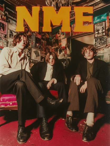 The image features three men sitting on a plush red couch against a vividly decorated wall covered with posters and flyers. The wall's eclectic design includes various sizes and colours of paper, creating a textured backdrop. Above the men, large, bold yellow letters spell out "NME." The men are dressed in casual attire, with one wearing a suit jacket, and they are positioned in a relaxed manner with legs crossed. The floor is carpeted in a bright red colour, complementing the couch. Overhead, a portion of an ornate ceiling and a chandelier can be seen, adding to the room's opulent feel.