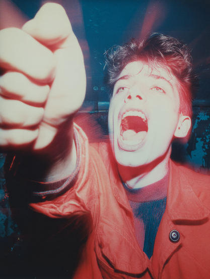 The image features a close-up of a person energetically gesturing with an outstretched fist toward the camera. The person has short, dark hair and an open-mouthed expression, suggesting they are either shouting or cheering. Vibrant red lighting illuminates their face and clothing, casting intense highlights and shadows. The individual is wearing a bright red jacket over a dark sweater, with a single large button visible on the lapel. The background appears to be dimly lit with blue tones, emphasising the vividness of the red lighting in the foreground.