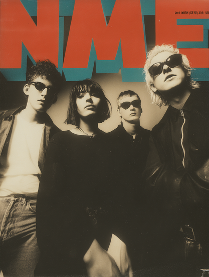 The image features a band of four individuals standing closely together, gazing directly at the camera. The photograph is tinted in sepia, creating a vintage effect. All members are wearing sunglasses, adding a sense of mystery. The individual on the left has curly hair and wears a white shirt under a jacket, standing with a casual posture. Next, a woman with shoulder-length dark hair wears a black top. Beside her stands a person with short, light hair in a light-coloured shirt. On the right, an individual with light hair wears a zip-up jacket. The bold red letters "NME" dominate the upper section of the image, set against a blue background.