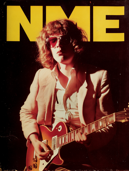 The image is a magazine cover featuring a person with curly hair, wearing large sunglasses and a light-coloured blazer over a button-up shirt. The individual is playing an electric guitar with a sunburst finish, positioned mid-frame. The background is dark, making the subject stand out under a warm, dramatic light that casts shadows. At the top, large yellow letters spell out "NME," taking up the upper portion of the cover.