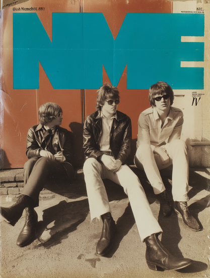 The image is a vintage magazine cover featuring three individuals sitting on a footpath against a large red metal door. They are dressed in stylish, retro clothing. On the left, one person wears dark pants, black boots, a white shirt, and a black leather jacket, with sunglasses and medium-length hair. The individual in the middle wears white pants, a black leather jacket, sunglasses, and has tousled medium-length hair. The person on the right, wearing light-coloured pants, a pale jacket, and sunglasses, also has similar length hair. Large blue letters "NME" dominate the top half of the image. The surface shows signs of wear, with visible creases and discolourations.