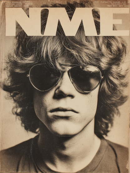 The image is a magazine cover featuring a close-up, black and white photograph of a person with shoulder-length wavy hair. The individual wears aviator sunglasses, and their expression is neutral, with lips slightly parted. The lighting casts gentle shadows, emphasising the texture of the hair and sunglasses. The top section of the cover prominently displays the large, bold letters "NME" in a blocky font, centred above the photograph. The overall tone of the image is vintage, with a slightly sepia hue that adds a retro feel.