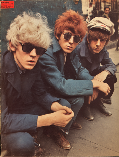 The image shows three young men sitting close together on an urban footpath. The person on the left has light blonde, voluminous hair and wears dark sunglasses, a dark shirt, and a blue jacket. The middle individual has bright red hair and round sunglasses, wearing a blue jacket with a white shirt underneath. The person on the right has brown hair and wears a cap, also dressed in a blue jacket. They are posed casually, with their knees bent and leaning slightly forward. The background features a blurred city street scene with a wrought iron fence and an indistinct figure in the distance, emphasising an urban environment.