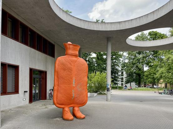 The image features a large, orange, hot water bottle sculpture with human-like legs, standing in an outdoor plaza. The sculpture is textured with ridges, resembling the surface of a real hot water bottle. It is positioned in front of a modern building with a concrete structure and circular openings in the ceiling, providing shade. The building has a glass entrance and windows with red frames. In the background, there are trees and a bicycle parked nearby. The plaza is paved with grey tiles, and…