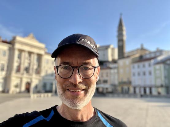 Me, taking a selfie at Piazza Tartini, Piran, slovenia. @lysander07 The image features a man standing in the foreground, taking a selfie. He is wearing a black cap with the word "HULDA" printed on it, a black t-shirt with blue accents, and glasses. The man has a gray beard and is smiling. In the background, there is a large, open square with classical architecture, including a prominent building with columns and a tall tower with a spire. The sky is clear and blue, indicating a sunny day. The b…