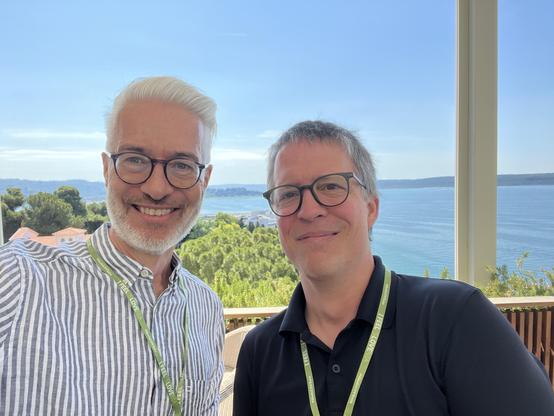 Me (left) and Axel, smiling for a selfi above the adriatic sea. @lysander07 The image features two men standing close together, smiling at the camera. They are both wearing glasses and lanyards with green tags. The man on the left has white hair and a beard, wearing a striped shirt, while the man on the right has gray hair and is wearing a black polo shirt. They are positioned in front of a large window that offers a scenic view of a body of water, likely a lake or sea, with lush green trees an…