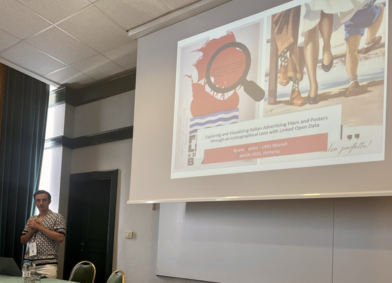 Bruno Sartini presenting his talk at the SemDH 2025 workshop. Bruno is standing behind a desk in front of the projection screen, showing the cover slide of his presentation, which has historical advertisement posters as a background.