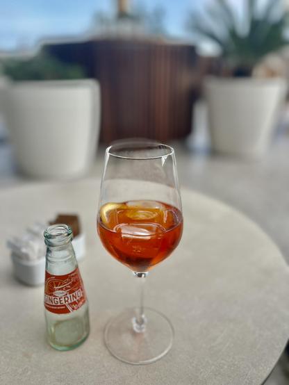 A wineglass filled with icecubes and an orange bitter lemonade, called "gingerino", standing on a white table next to a small and empty bottle of Gingerino. The rest of the scenery is the inventory of a roof terrace, all out of focus of the camera.