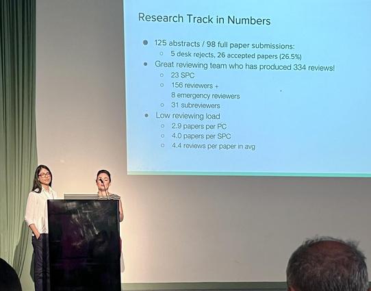 The image shows a presentation slide titled "Research Track in Numbers" displayed on a projector screen. The slide details the statistics of a research track, including the number of abstracts and full paper submissions, the acceptance rate, and the reviewing team's performance. It states that there were 125 abstracts and 98 full paper submissions, with 5 desk rejects and 26 accepted papers, representing a 26.5% acceptance rate. The reviewing team consisted of 23 Senior Program Committee (SPC) …