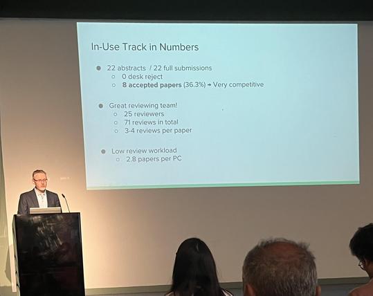The image shows a presentation slide titled "In-Use Track in Numbers" displayed on a large screen. The slide contains bullet points with statistical information about the track. The first bullet point states that there were 22 abstracts and 22 full submissions, with 0 desk rejects and 8 accepted papers, which is 36.3% of the submissions, indicating a very competitive track. The second bullet point highlights the "Great reviewing team!" with 25 reviewers, 71 reviews in total, and an average of 3…