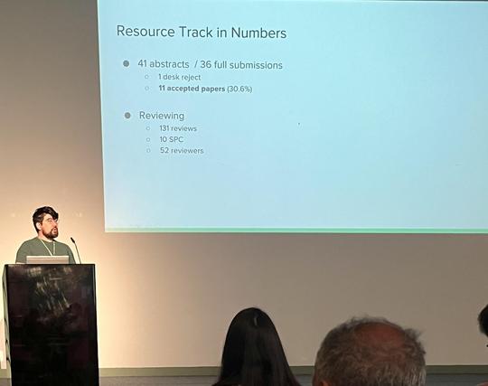 The image shows a presentation slide titled "Resource Track in Numbers" displayed on a large screen. The slide contains bullet points with numerical data. The first bullet point states "41 abstracts / 36 full submissions," with sub-points indicating "1 desk reject" and "11 accepted papers (30.6%)." The second bullet point is labeled "Reviewing" and includes "131 reviews," "10 SPC," and "52 reviewers." A person is standing at a podium to the left of the screen, wearing a green shirt and a lanyar…