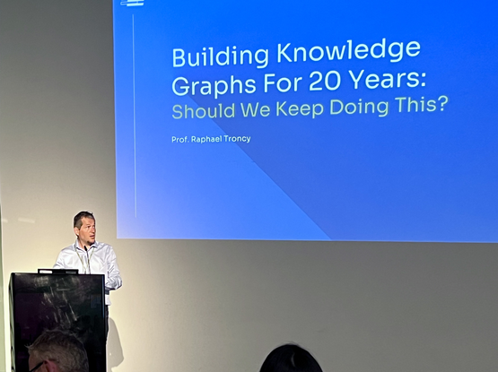 The image shows a presentation setting with a speaker standing at a podium. The speaker is a man wearing a light-colored shirt and a lanyard, addressing an audience. Behind him, a large screen displays a slide with a blue background and white text. The slide's title reads "Building Knowledge Graphs For 20 Years: Should We Keep Doing This?" and includes the name "Prof. Raphael Troncy" below the title. The presentation appears to be taking place in a conference or seminar room, with a plain wall …