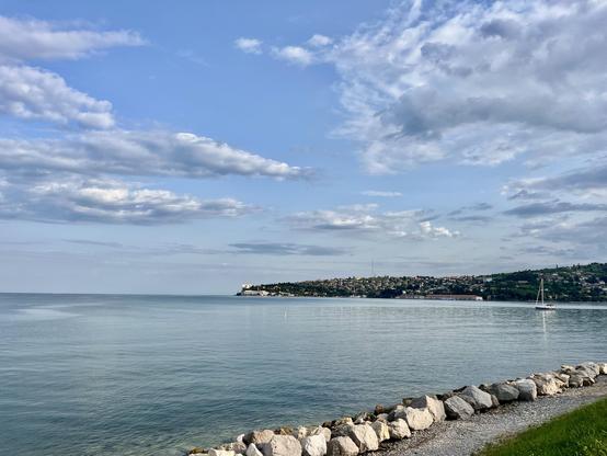 Portoroz seaside view. @lysander07 The image depicts a serene coastal scene. In the foreground, a rocky shoreline with large, irregularly shaped stones borders a calm, expansive body of water. The water is a tranquil blue, reflecting the partly cloudy sky above. A small white sailboat is anchored in the distance, adding a touch of life to the scene. The background features a hilly landscape with a densely populated area, likely a town or city, characterized by numerous buildings and greenery. T…