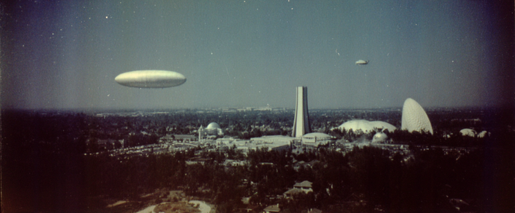 A futuristic landscape with domed and cylindrical buildings beneath a clear sky featuring two zeppelin-like airships.