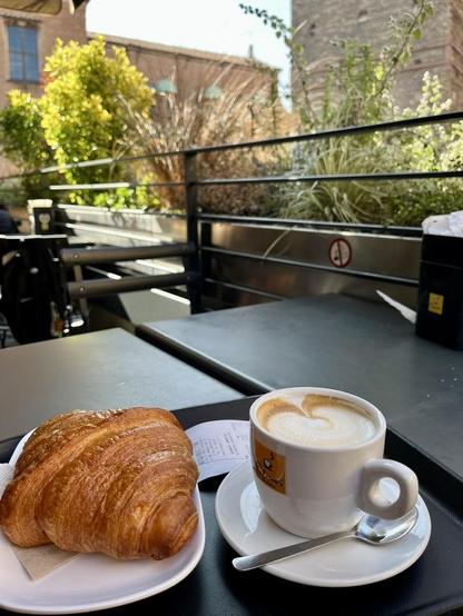 A cup of Cappuccino with an "empty Croissant" (Cornetto vuotuto) on a small plate, both next to each other on a dark table placed outside on a roof terrace close to the "Due Torre" (Two Towers) in Bologna city center. 