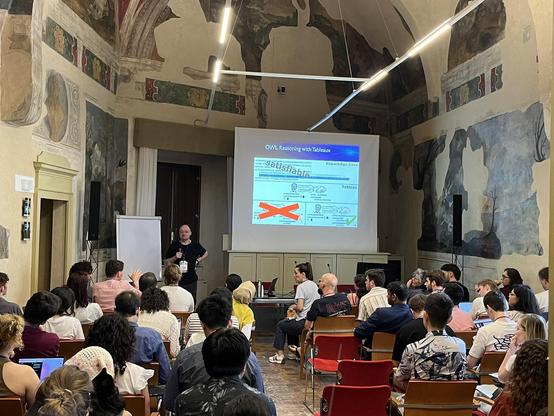 Sebastian Rudolph standing next to the presentation screen with the penultimate slide of his presentation. The audience is paying attention to him answering students' questions.