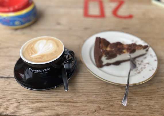 A dark brown cup of Cappuccino. The Cappuccino shows some Latté-Art in the form of a heart. Next, a piece of Zupfkuchen on a plate with a fork beside. In the upper left corner of the picture, we see the fragment of a Tunesian style ceramic ash tray (very colorful). On the table surface (mace of wood) there is the number 20 (as a helper for the service, when you are ordering your drink/cake inside)