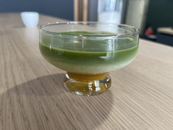 Mango Matcha Latte, i.e. a glass with Mango/apple sauce on the bottom, then vanilla oat milk in the middle, topped with gree matcha. The glass is standing in the middle of a long counter made of wood. The background is blurred.
