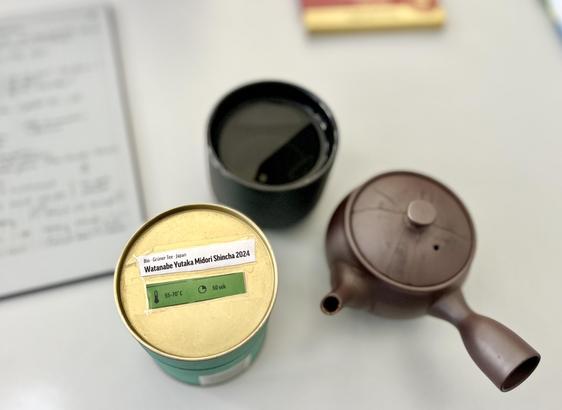 A box of tea with a sticker on the top saying" Bio Green Tea - Japan - Watanabe Yukata Midori Shincha 2024", next to a black cup of tea and a single hand tea pot. All three objects are placed on top of a grey desktop, photographed from the top. On the left in the beokeh, we can see a "remarkable" tablet with text written on it (which cannot be deciphered).