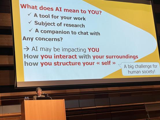Yuko Harayama standing at the speakers desk in fron of her projected slides.
First slide:
What does AI mean to you?
- a tool for your work
- subject of research
- a companion to chat with
Any concerns?
-> AI may be impacting YOU
How you interact with your surroundings
How you structure your "self" -> a challenge for human society