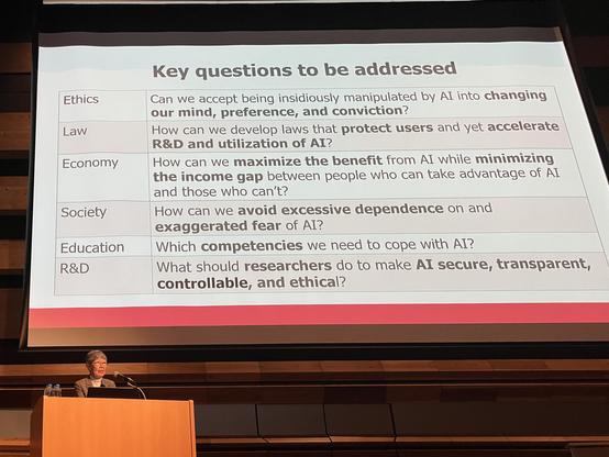 Yuko Harayama standing at the speakers desk in front of her projected slides.
2nd slide:
Key Questions to be addressed:
- Ethics: Can we accept being insiduously manipulated by AI into change our mind, preference, and conviction?
- Law: How can we develop law that protect users and yet accellerate R&D and utilization of AI?
- Economy: How can we maximize the benefit from AI while minimizing the income gap between people who can. take advantage of AI and those who can't?
- Society: How can we a…