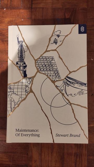 A book lying on a wooden table. It's titled Maintenance: of Everything and it's by Stewart Brand. The cover shows diagrams of various things that require maintenance such as the statue of liberty, a writing system and a ship at sea. Each is drawn within a shard, and the shards are outlined with gold, giving the impression of a Kintsugi piece of art.