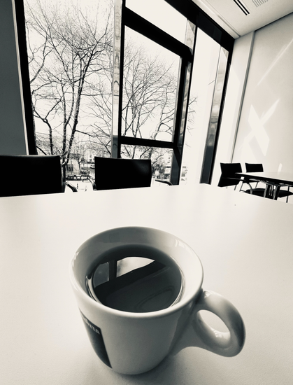 black/white photograph of a cup of tea standing in the foreground on a white table, surrounded by chairs. Large office windows in the background with trees standing directly behind the windows. The trees are still without leaves. It is early March...