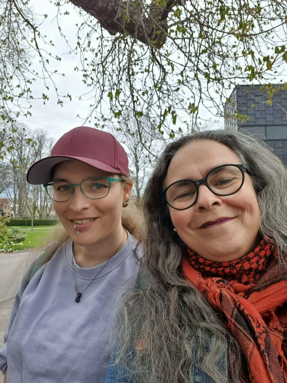 Two people with a background of spring. I'm wearing a light purple sweater and red cap. The other slightly in front of me wearing a red/black scarf and both wearing glasses.