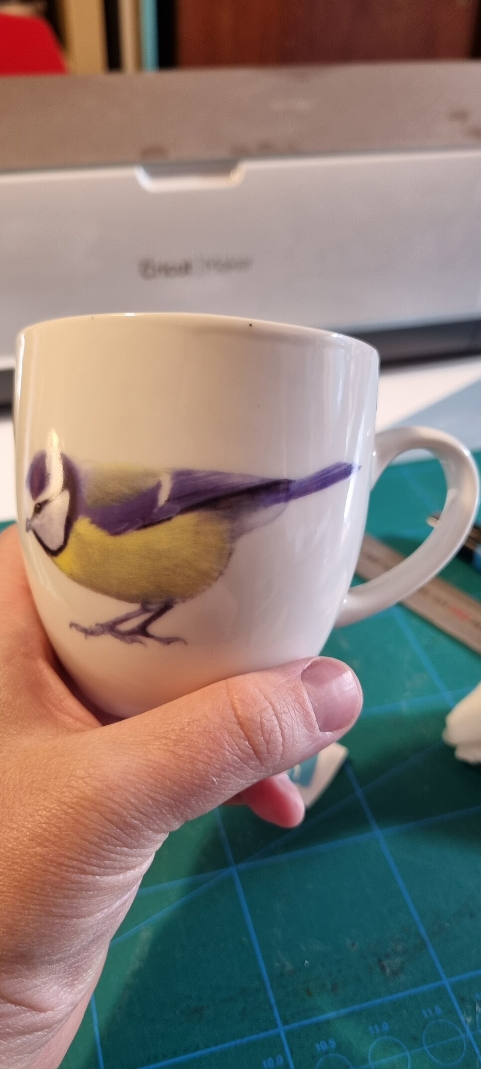 My hand holding a round cup with a picture of a blue tit on it. In the background you can see an out of focus cricut machine