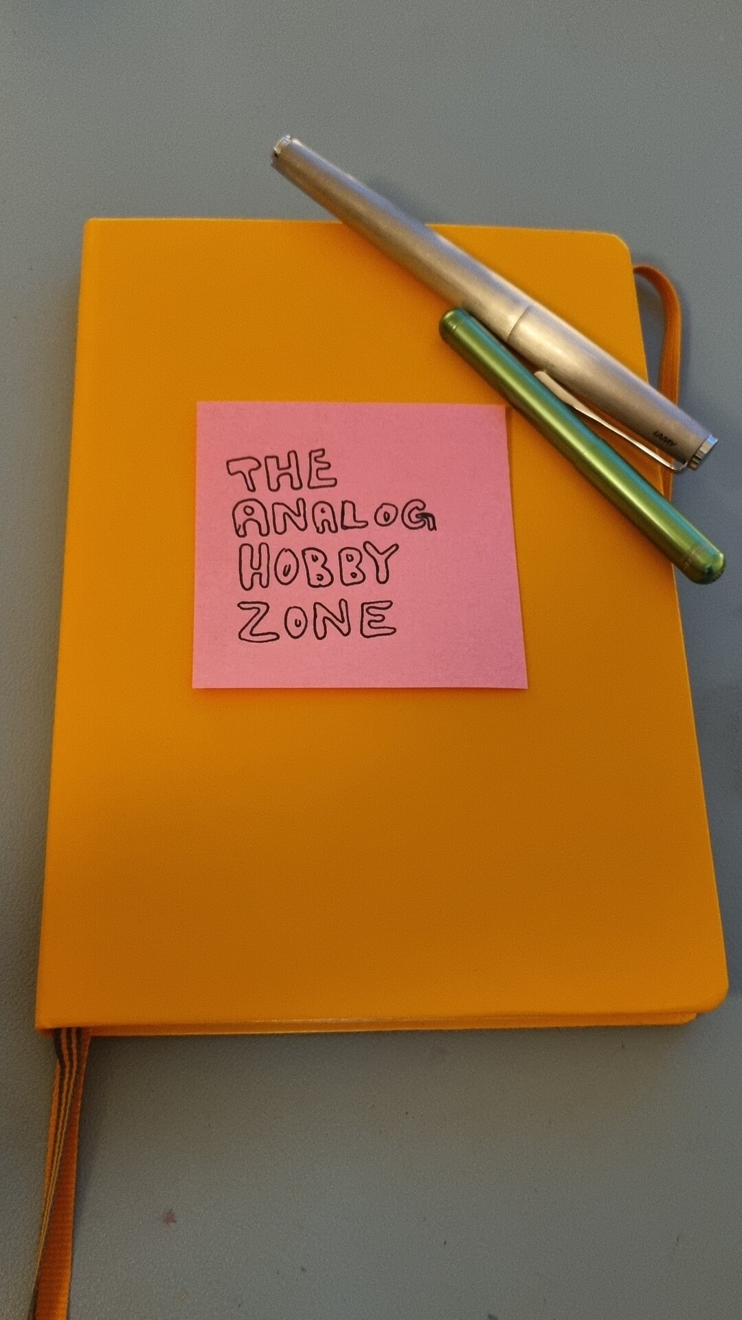 A pink postit style note on an orange book. Black block letters on the note say "THE ANALOGNHOBBY ZONE". There are two pens there too.