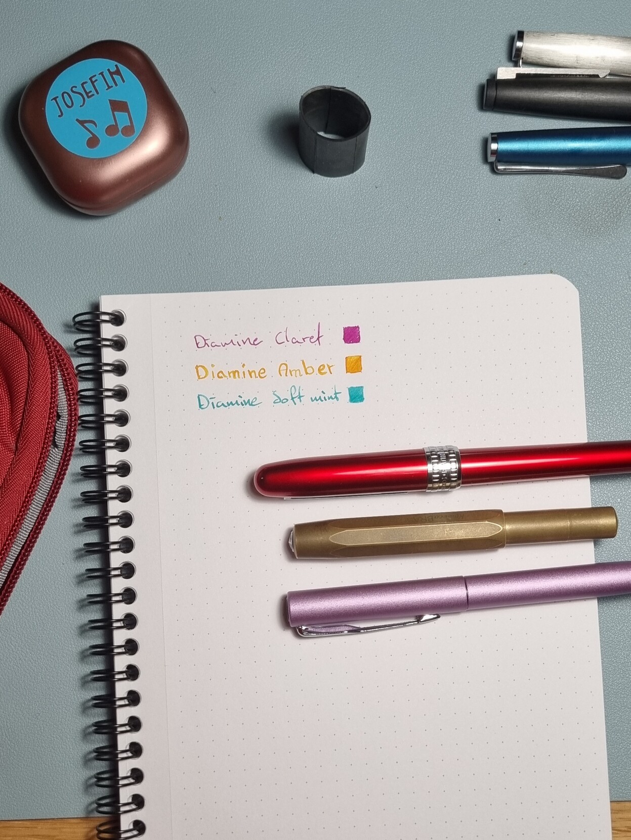 A somewhat arranged picrure of a notepad with some thinga strewn around. On the note pad it says "Diamine Claret" "Diamine Amber" and "Diamine Soft mint". There are three pens coresponding to the different inks.