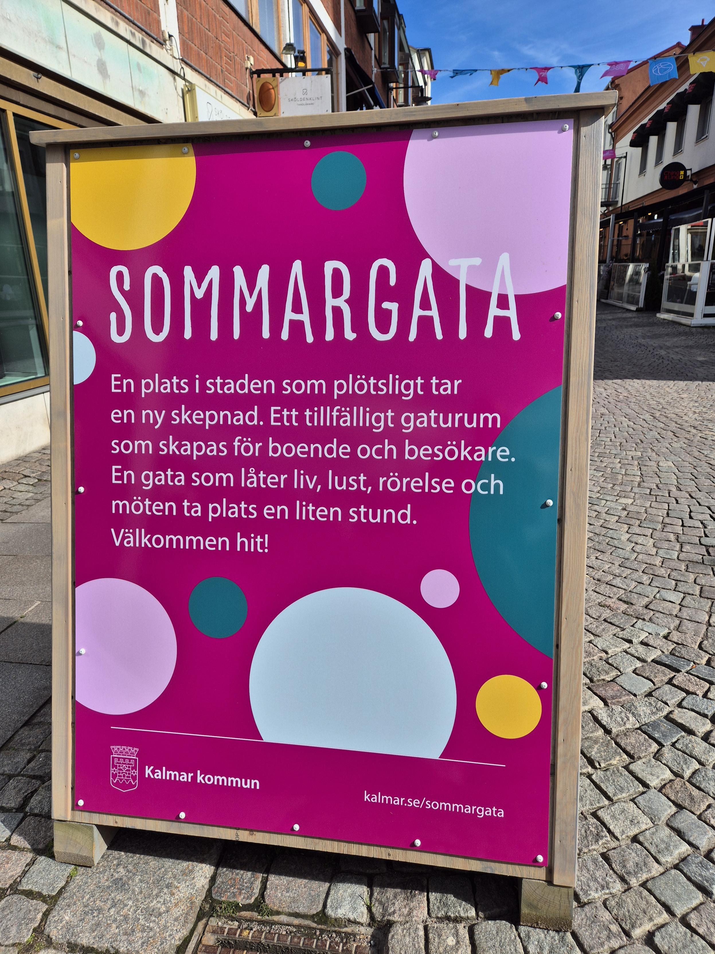 En skylt med texten: Sommargata. En plats i staden som plötsligt tar en ny skepnad. Ett tillfälligt gaturum som skapas för boende ich besökare. En gata som låter liv, lust, rörelse och möten ta plats  en liten stund. Välkommen hit.
