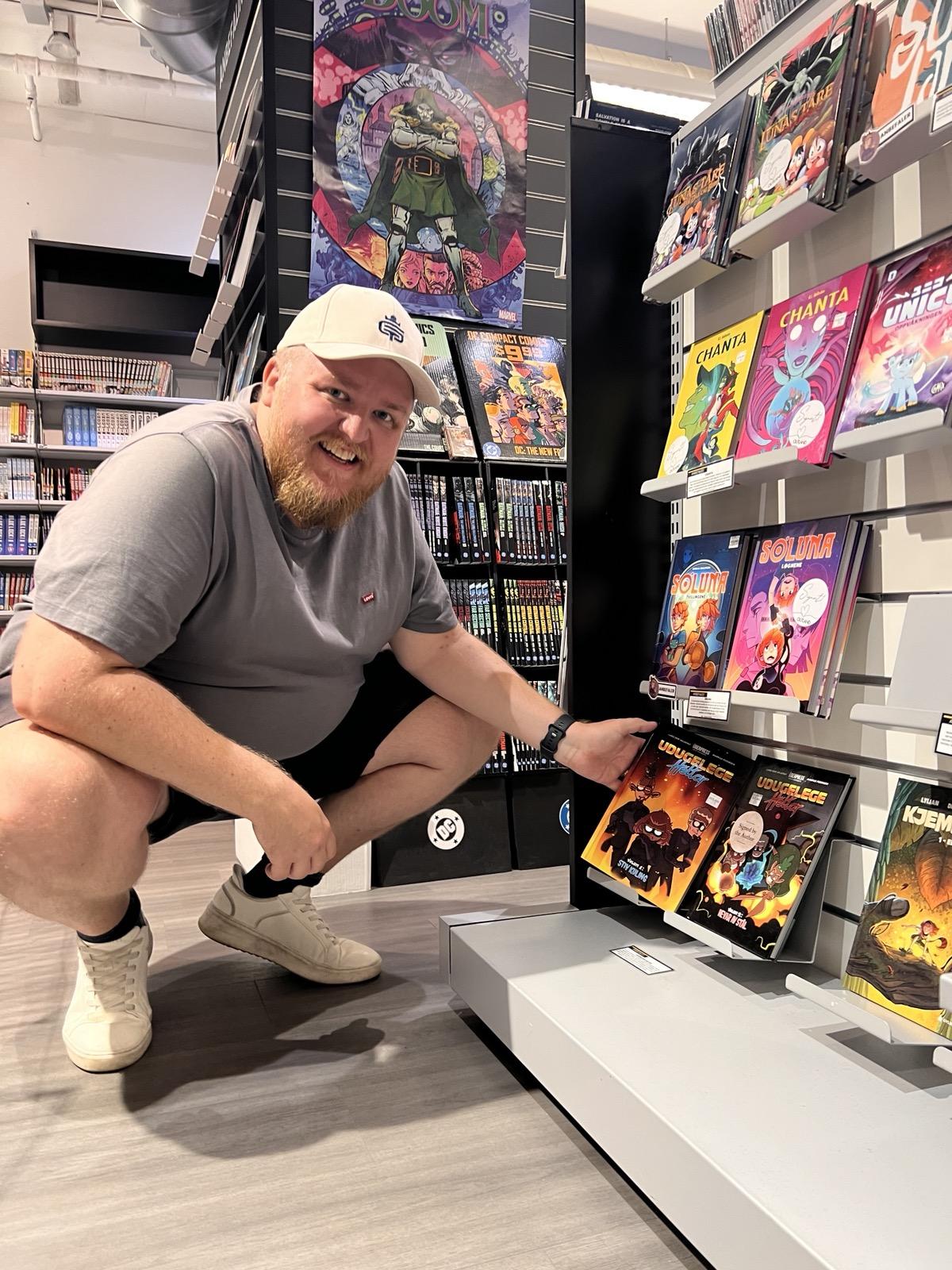 Eit innendørsbilete frå butikken Outland i Stavanger. Ein smilande mann med skjegg, ljos caps, grå t-skjorte, mørk shorts og kvite joggesko sit på huke ved ei hylleseksjon. Han peikar stolt mot to teikneseriebøker som står utstilte nedst på hylla: Udugelege heltar: Volum 1 – Stiv kuling og Udugelege heltar: Volum 2 – Nevar av stål. Hyllene rundt er fylte med ulike teikneseriar og grafiske romanar som Chanta og Soluna, og i bakgrunnen heng det ein plakat med Marvel-figuren Doctor Doom på ein reol med fleire teikneserieutgåver.